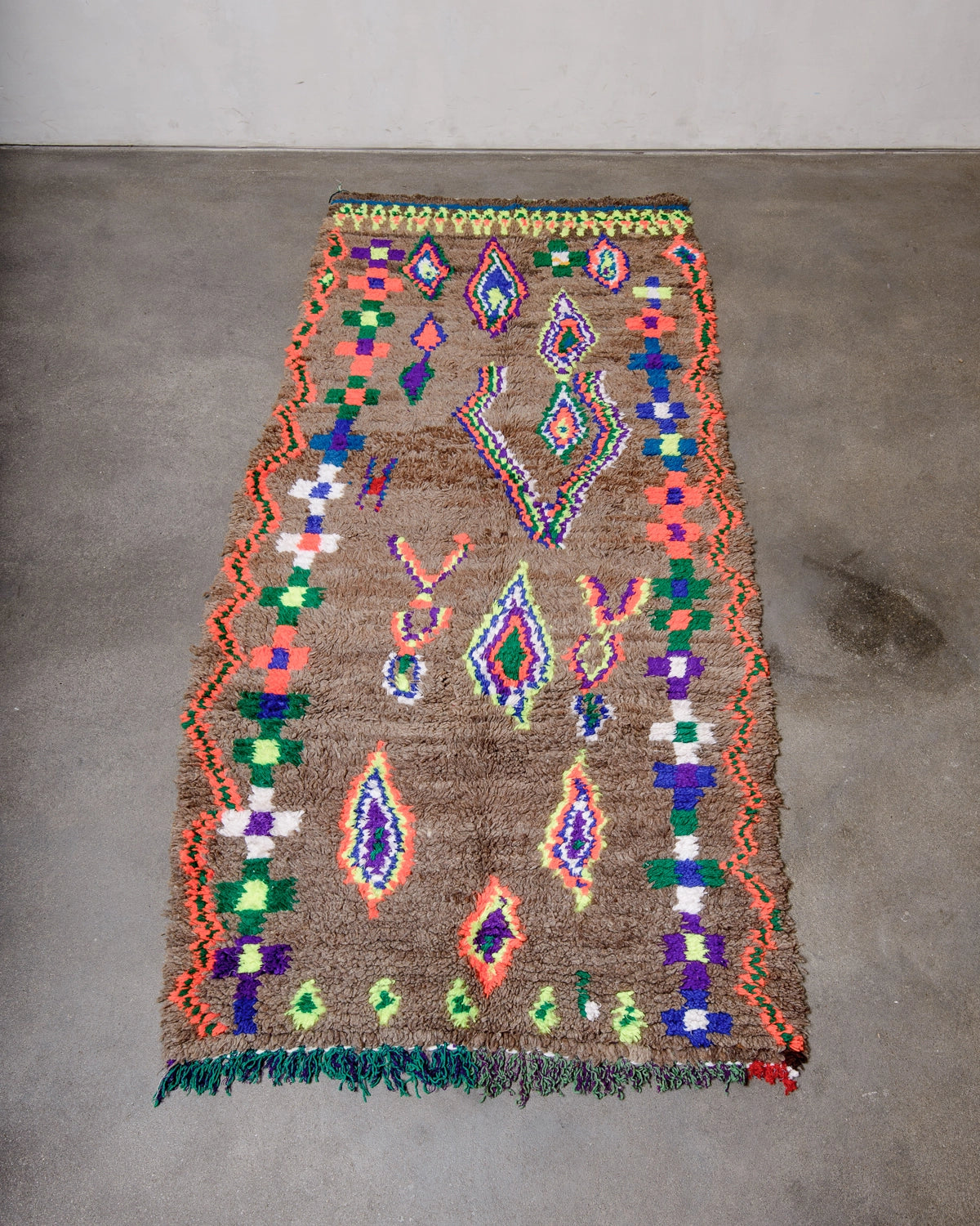 Vintage Berber Rug made of brown wool with design details in neon, purple, blue, green cotton, laying on concrete floor.